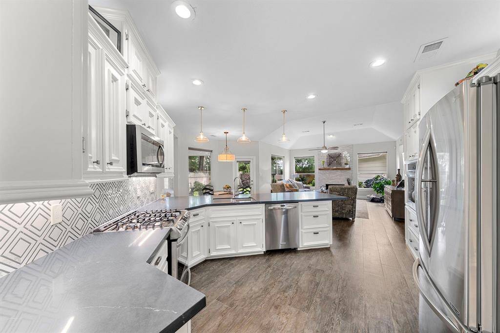 This photo showcases a modern, open-concept kitchen with sleek stainless steel appliances, white cabinetry, and a stylish geometric backsplash. The space features a large island with a built-in sink and dishwasher, offering ample counter space. The kitchen flows seamlessly into a cozy living area, highlighted by wooden floors and plenty of natural light from surrounding windows.