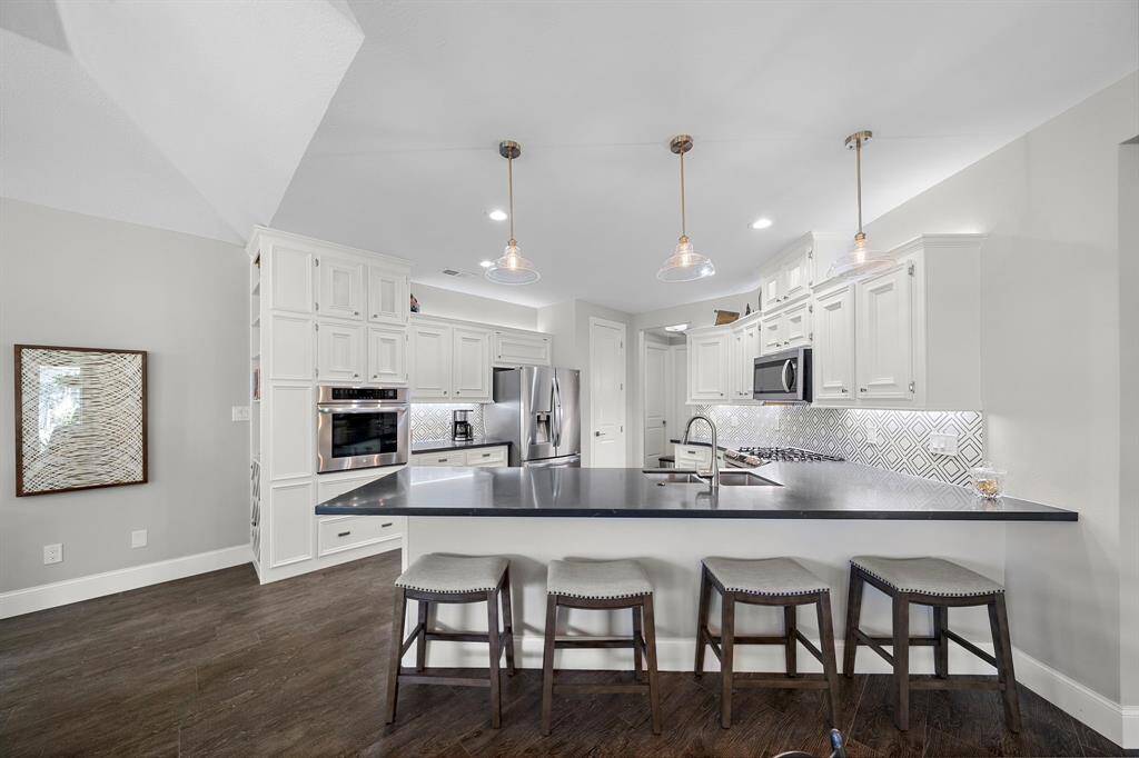 This modern kitchen features sleek white cabinetry, a spacious dark countertop, and stylish pendant lighting. It includes stainless steel appliances, a geometric backsplash, and a breakfast bar with seating for four, perfect for entertaining.