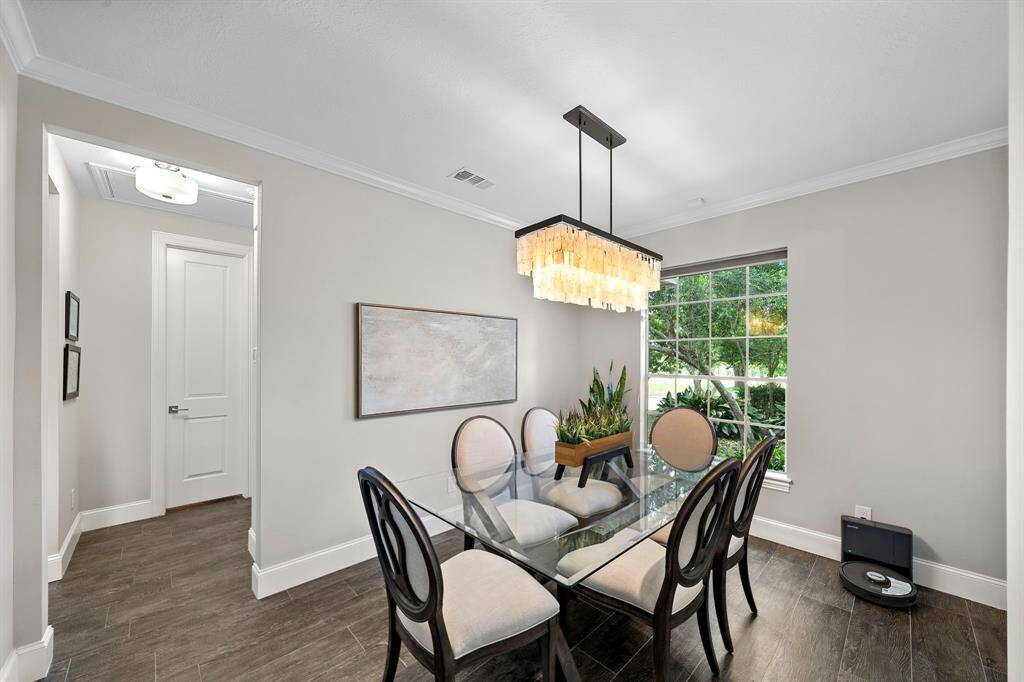 This dining area features a sleek glass table with elegant chairs, modern chandelier, and large window providing natural light. The neutral walls and dark wood flooring create a warm, inviting atmosphere.