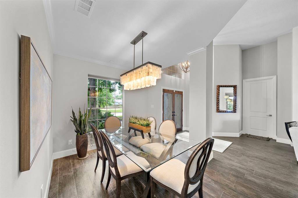 This elegant dining area features a modern chandelier, glass-top table, and stylish chairs. The space is brightened by a large window and complemented with tasteful decor and wood flooring, offering a welcoming and contemporary atmosphere.