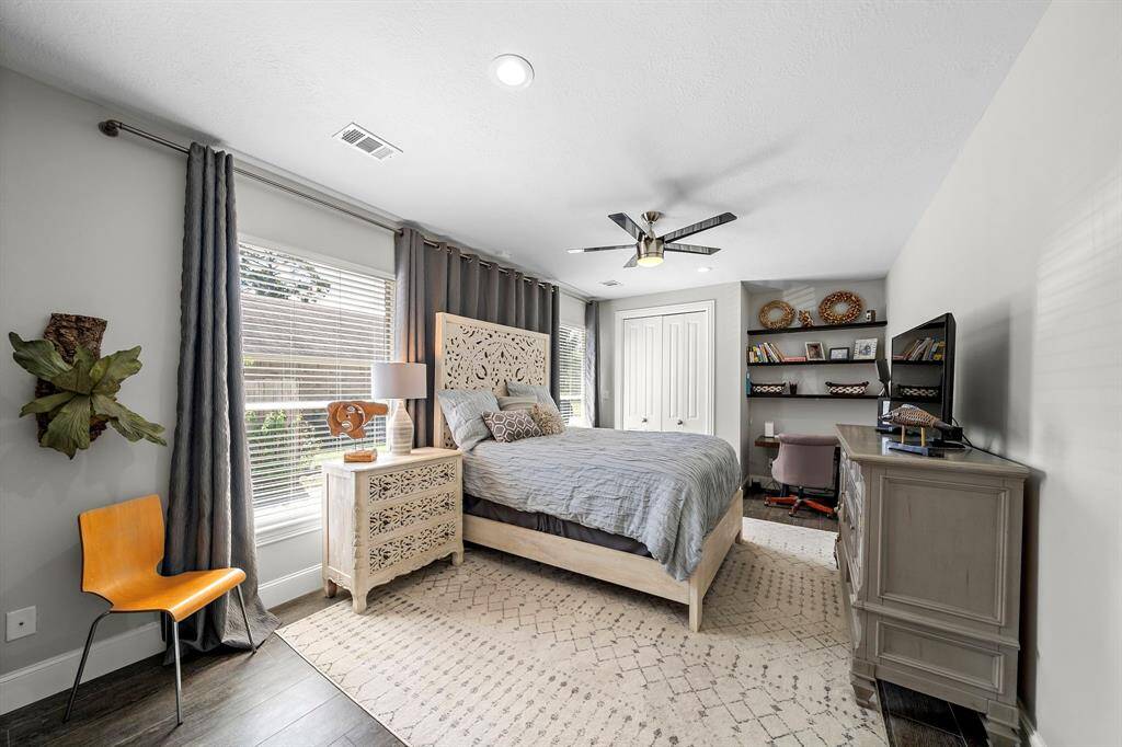 This bright bedroom features a stylish bed with an intricate headboard, a cozy workspace with shelves, and a large window with elegant drapes. The room is accented with a modern ceiling fan and tasteful decor, creating a welcoming and functional space.