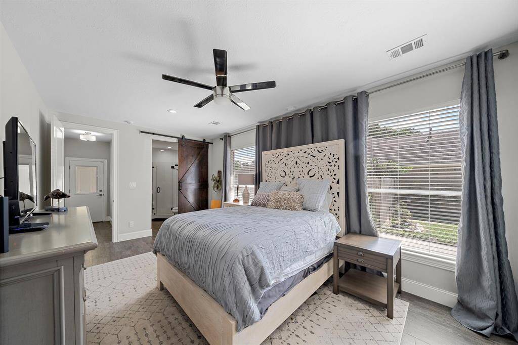 This bedroom features a modern design with a decorative headboard and neutral tones. It includes large windows for natural light, a ceiling fan, and a sliding barn door leading to the bathroom. The space is inviting and functional, with ample room for furniture.