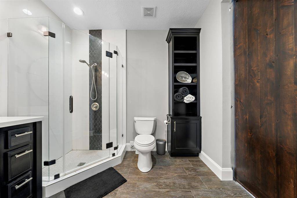 This bathroom features a modern glass shower with sleek fixtures, a white toilet, and ample storage with dark cabinetry. The space is complemented by wood-look tile flooring and a stylish wood accent wall.