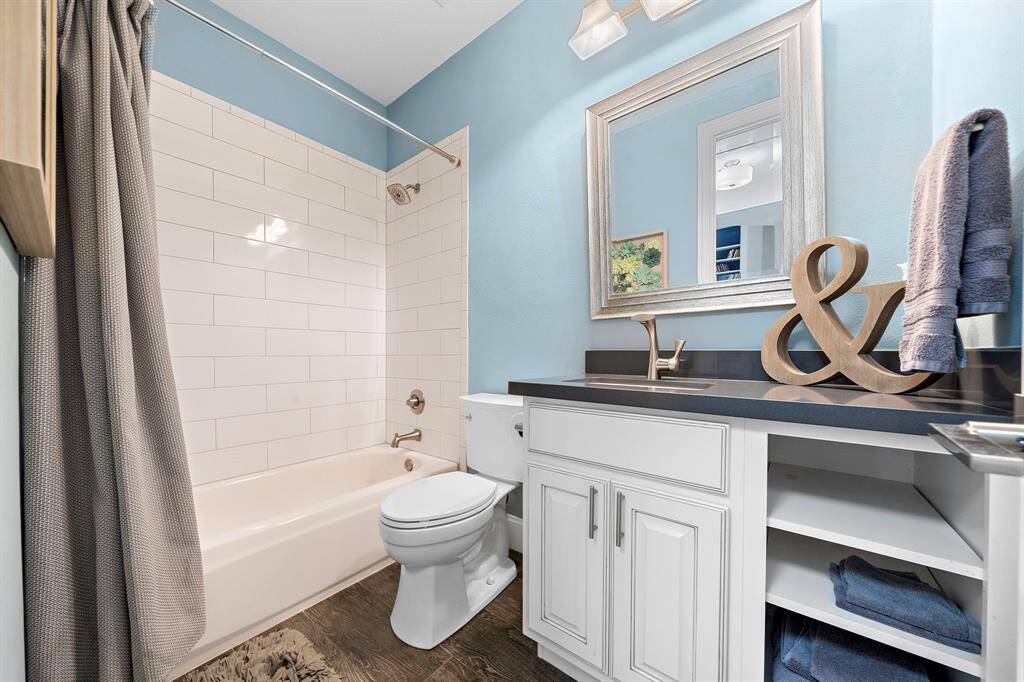 This bathroom features a modern design with light blue walls and a white subway-tiled shower. It includes a sleek black countertop, white cabinetry, and a large framed mirror. The space is complemented by wood accents and a neutral shower curtain.