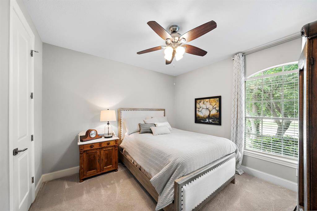 Cozy bedroom with neutral tones and a large window providing plenty of natural light. Features a ceiling fan, wooden bedside table with a lamp, and tasteful wall art.