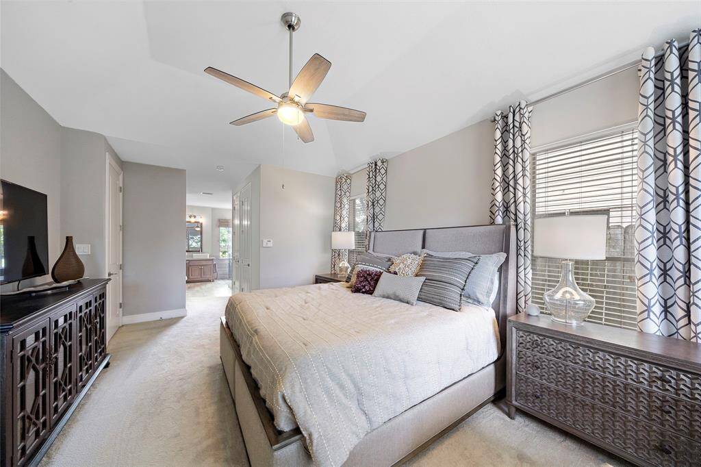 This photo showcases a spacious, well-lit bedroom with modern decor. It features a large bed with plush bedding, stylish nightstands, and patterned curtains. A ceiling fan and neutral tones create a cozy atmosphere. The room connects to an en-suite bathroom, visible through an open doorway.