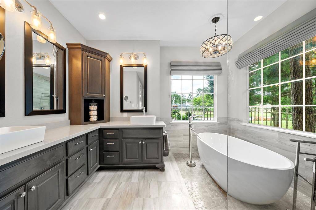 This elegant bathroom features a freestanding tub with a large window view, dual vessel sinks on a spacious vanity with ample storage, and stylish lighting fixtures, creating a bright and inviting space.