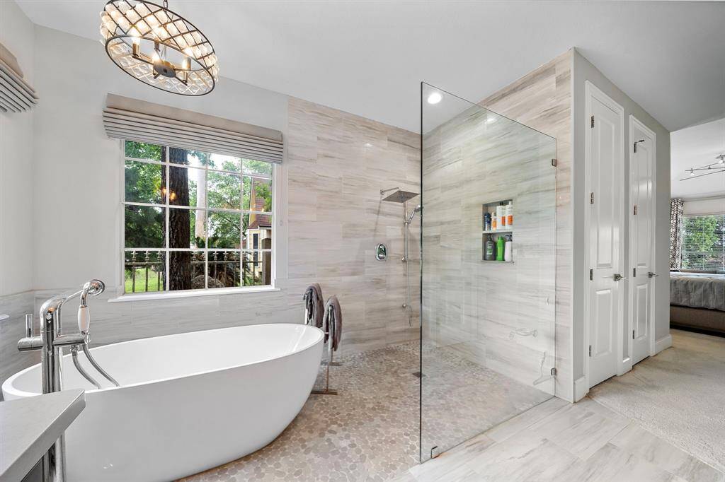 This bathroom features a modern design with a freestanding tub, glass-enclosed shower, and large window providing natural light. Neutral tones and sleek finishes create a relaxing atmosphere, seamlessly connecting to the adjacent bedroom.