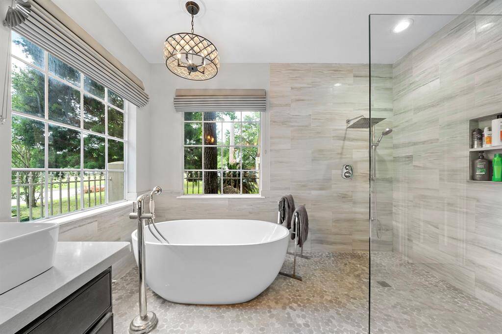 This modern bathroom features a freestanding bathtub with a floor-mounted faucet, a spacious glass-enclosed shower, and large windows offering natural light and views of greenery. A stylish light fixture enhances the elegant design.