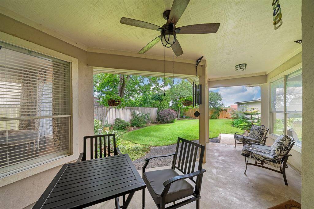 This photo showcases a cozy covered patio with a ceiling fan, outdoor seating, and table. It overlooks a well-maintained, fenced backyard with lush greenery, providing a perfect space for relaxation and entertaining.
