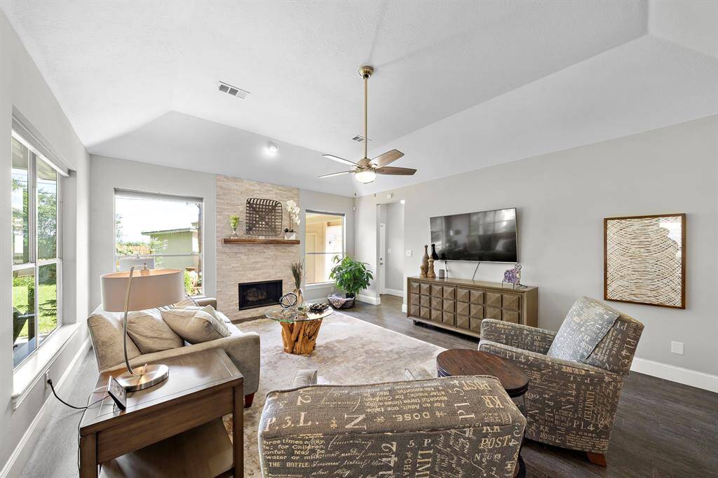 This inviting living room features a cozy fireplace, large windows for natural light, and modern decor. It has a comfortable seating area, sleek hardwood floors, a ceiling fan, and a mounted TV, creating a perfect space for relaxation and entertainment.
