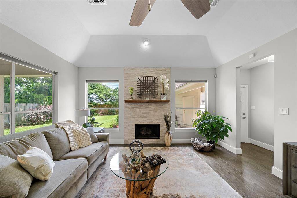This living room features a high ceiling, modern ceiling fan, and a cozy fireplace with a stone accent wall. Large windows offer abundant natural light and views of the landscaped backyard. The space is elegantly furnished with a neutral-toned sofa and stylish decor, creating a warm and inviting atmosphere.
