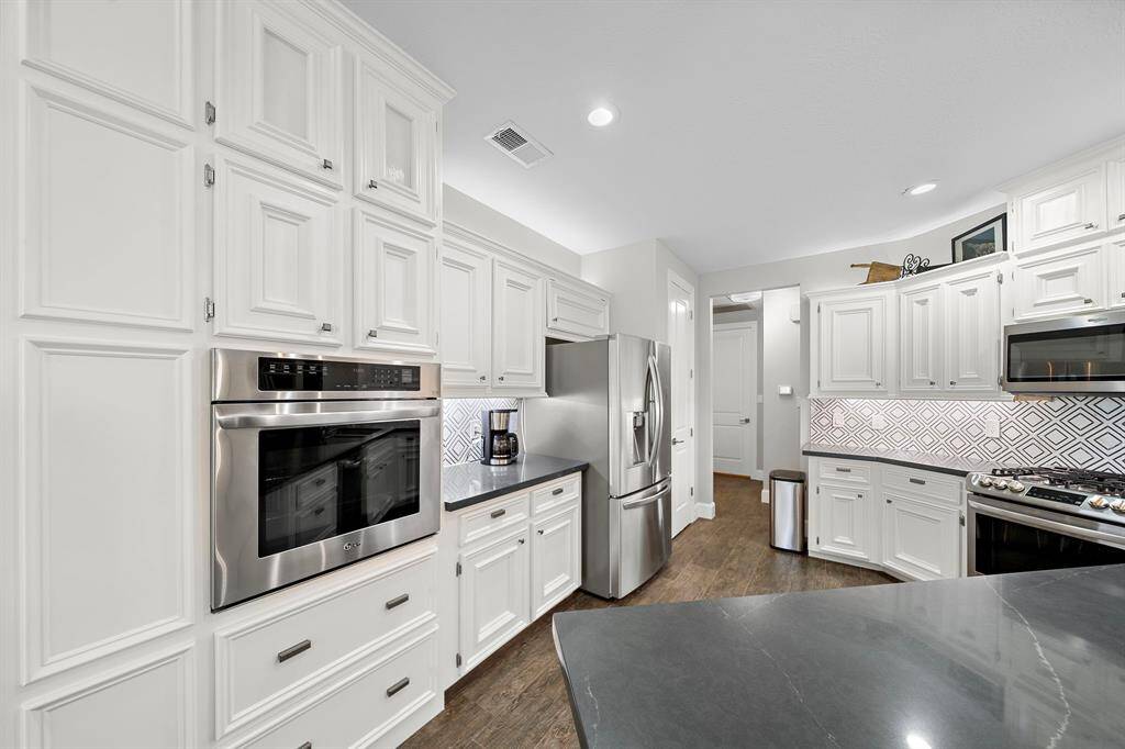 This kitchen features elegant white cabinetry, modern stainless steel appliances, and a stylish geometric backsplash. The space is well-lit with recessed lighting and has dark wood tile flooring, creating a sleek and inviting atmosphere.