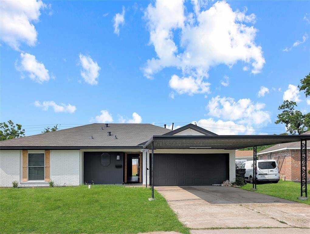This photo shows a single-story home with a modern exterior, featuring a dark garage door and trim. It has a well-maintained front lawn, a paved driveway, and a carport for additional parking. The house is accented with light brick and wood shutters, offering a clean and inviting curb appeal.