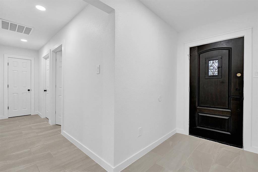 This photo shows a modern, bright entryway with a sleek, dark front door featuring a decorative glass panel. The space has light-colored tile flooring and white walls, creating a clean and inviting atmosphere.