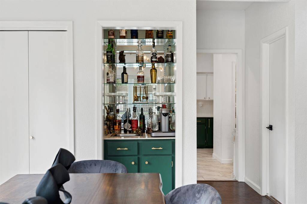 The dining room features a modern chandelier, a sleek wooden table with stylish chairs, and a built-in bar area with green cabinets. Large windows with elegant curtains allow natural light, and decorative elements add a contemporary touch.