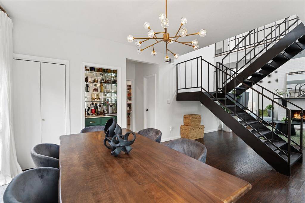 This photo showcases a stylish and modern open-plan living area with a sleek metal staircase, laminate floors, and a cozy fireplace. Large windows provide abundant natural light, and a chic chandelier adds elegance above the dining area. Perfect for comfortable living and entertaining.