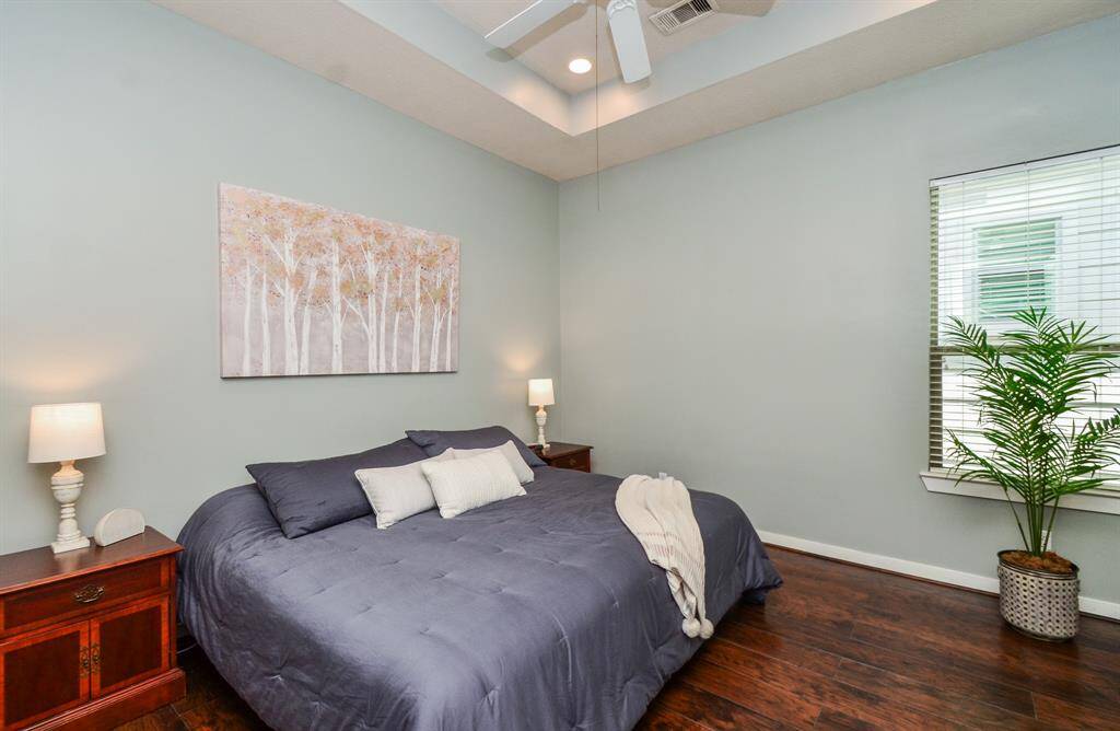 Huge Primary bedroom features natural walls, a tray ceiling with a fan, and warm wood flooring.  A window with blinds, and a decorative plant, creating a peaceful and inviting space.