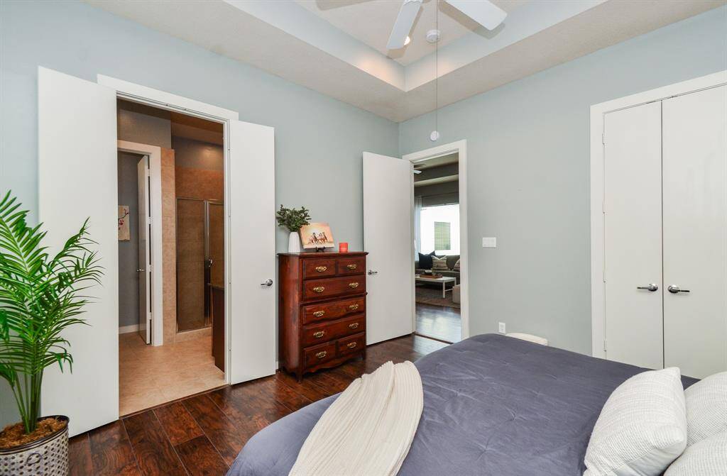 This bedroom photo features opened doors, hardwood floors, a ceiling fan, and neutral walls. It includes an ensuite bathroom, a closet with double doors, and a view into a cozy living area.