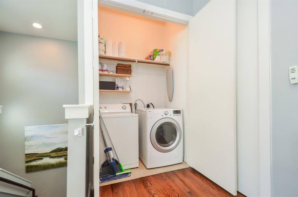 A compact laundry nook with a washer and dryer, shelving for storage, and neatly organized cleaning supplies. It features wood flooring and is adjacent to a staircase, providing a practical and efficient use of space.