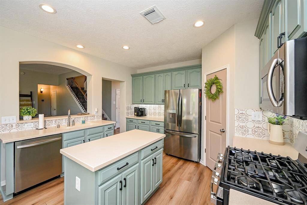 This kitchen features modern green cabinets, stainless steel appliances, and a stylish backsplash. It has an island, wood look vinyl flooring, and an open view to the adjacent room, offering a modern and inviting space.