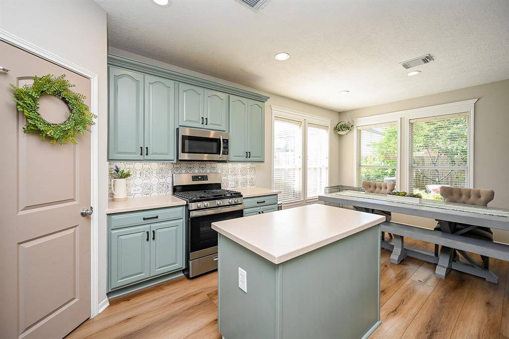 This bright kitchen features green cabinets, stainless steel appliances, and a central island. It opens to a cozy dining area with large windows providing ample natural light, set against warm wood look vinyl flooring.