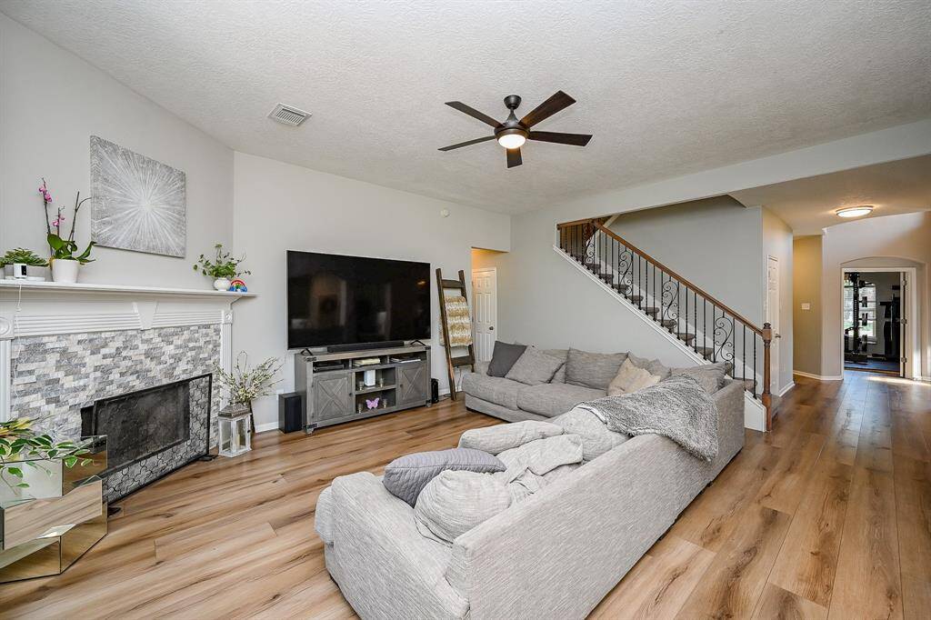 Spacious living room featuring a cozy fireplace and stylish ceiling fan. The room is accented by light wood look flooring and a wrought iron staircase, creating a welcoming and contemporary atmosphere.