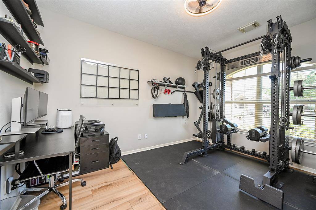 This photo showcases space for a versatile space currently used as a home gym and office space. organized workspace The room is bright, thanks to a large window allowing natural light, and has light wood look flooring.