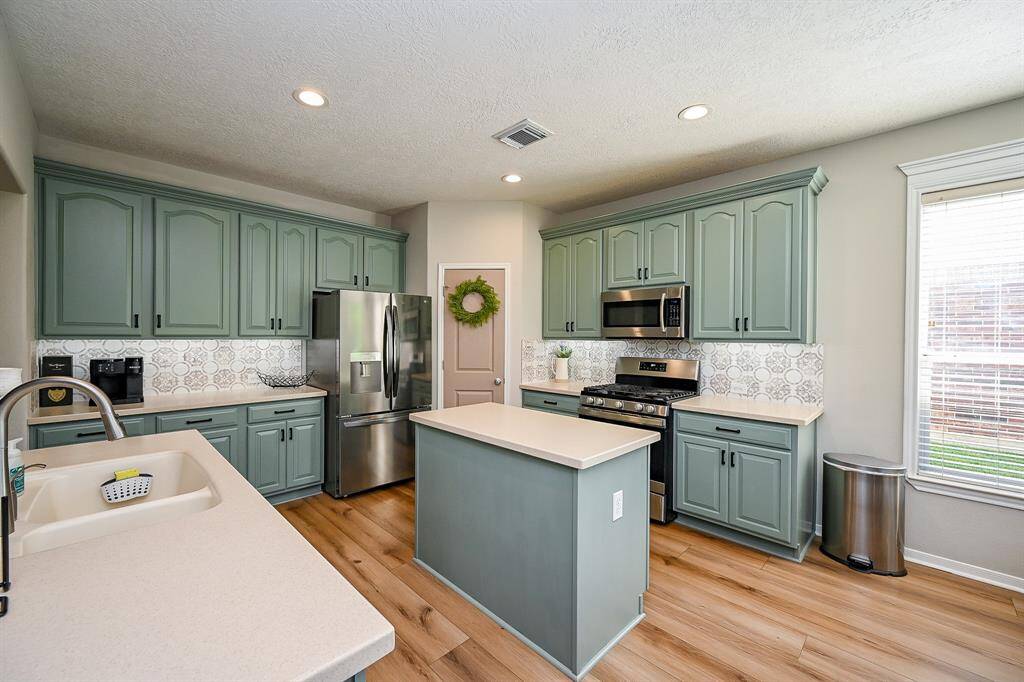 This kitchen features stylish green cabinetry, stainless steel appliances, and a central island. The light wood flooring and ample natural light create a warm, inviting atmosphere, while the backsplash adds a touch of elegance.