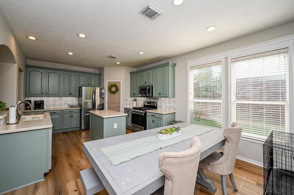 This kitchen features modern green cabinetry, stainless steel appliances, and a central island. It opens to a cozy dining area with ample natural light from large windows. The space combines style and functionality, perfect for family gatherings.