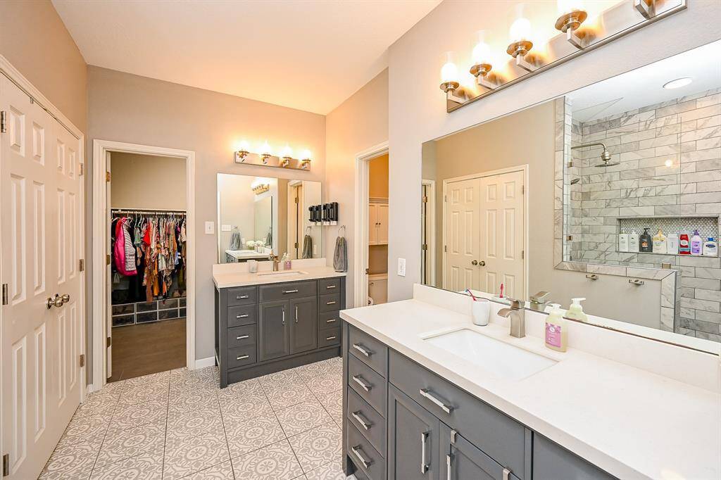 This spacious bathroom features dual sinks with modern fixtures, elegant tile flooring, and ample lighting. It includes a walk-in closet and a large, tiled shower offering a luxurious and functional design.