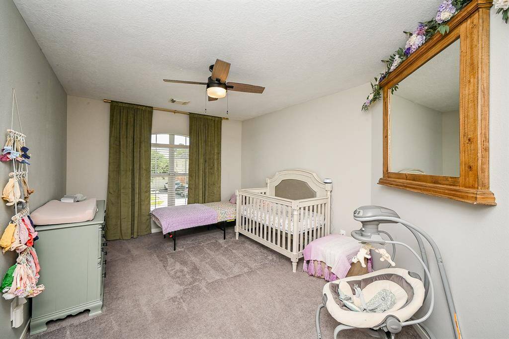 This cozy nursery features soft carpet, natural light streaming through large windows with green curtains. A ceiling fan adds comfort.