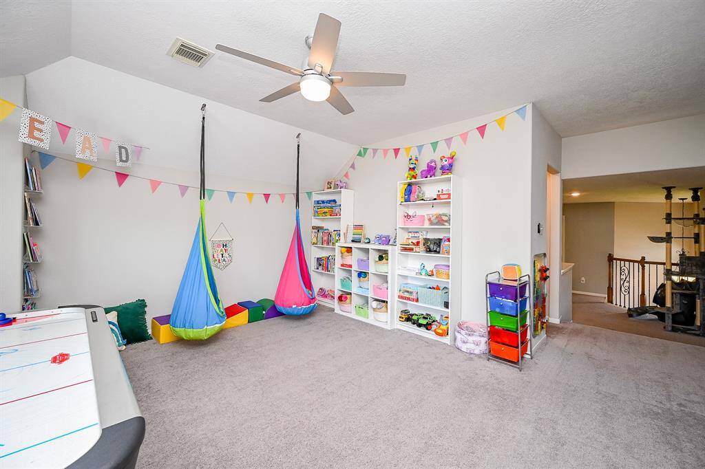 This spacious playroom features soft carpeting, and ample storage with shelves for toys and books. It includes a ceiling fan, creating a bright and inviting space perfect for many options.