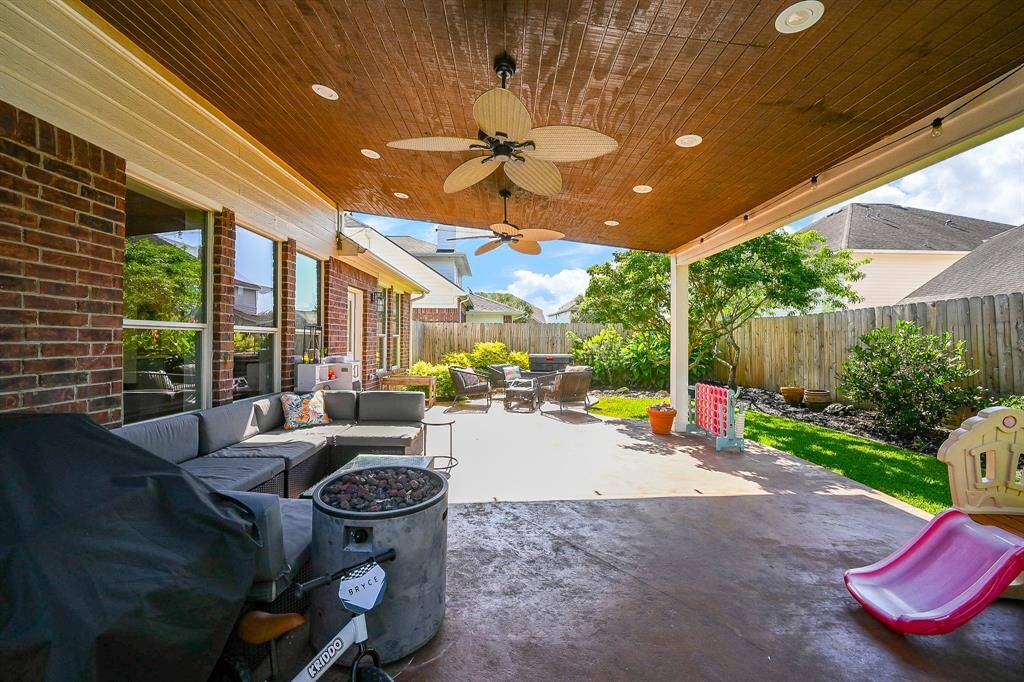 This inviting backyard features a spacious covered patio with a ceiling fan, perfect for relaxation. The yard is fenced, with well-maintained landscaping and a play area.