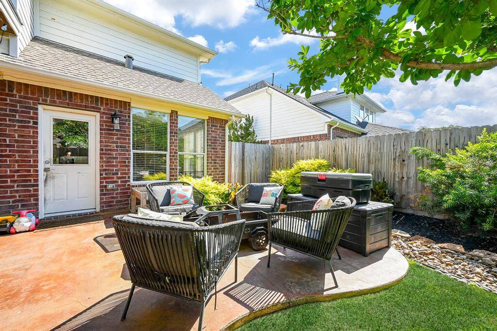 This photo shows a cozy backyard patio with, surrounded by a well-maintained garden and privacy fence. A brick exterior and large windows provide a charming backdrop. Perfect for relaxing and outdoor gatherings.