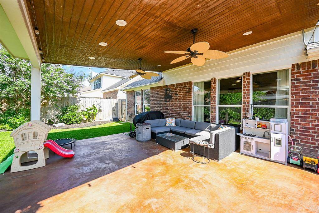 This outdoor space features a spacious covered patio with ceiling fans, perfect for relaxation. It includes a plenty of space for a cozy seating area, children's play equipment, and a small outdoor kitchen setup, all overlooking a well-maintained lawn. Ideal for family gatherings and entertaining.