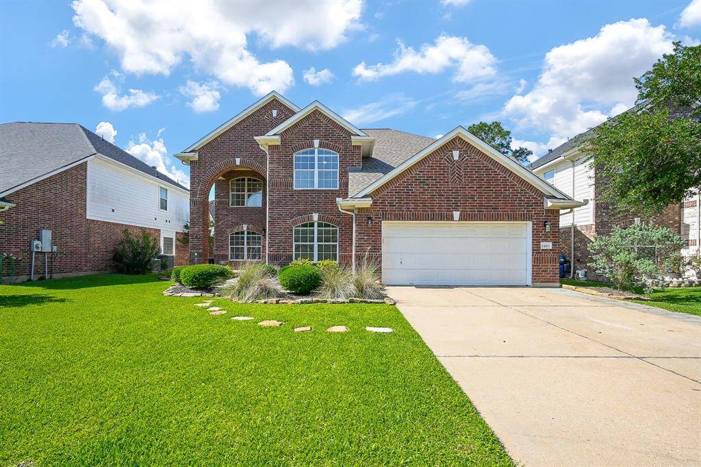 This is a charming two-story brick home with a spacious front yard and well-maintained landscaping. It features a two-car garage and a welcoming arched entryway. The home is set against a backdrop of blue skies and fluffy clouds, creating an inviting atmosphere.