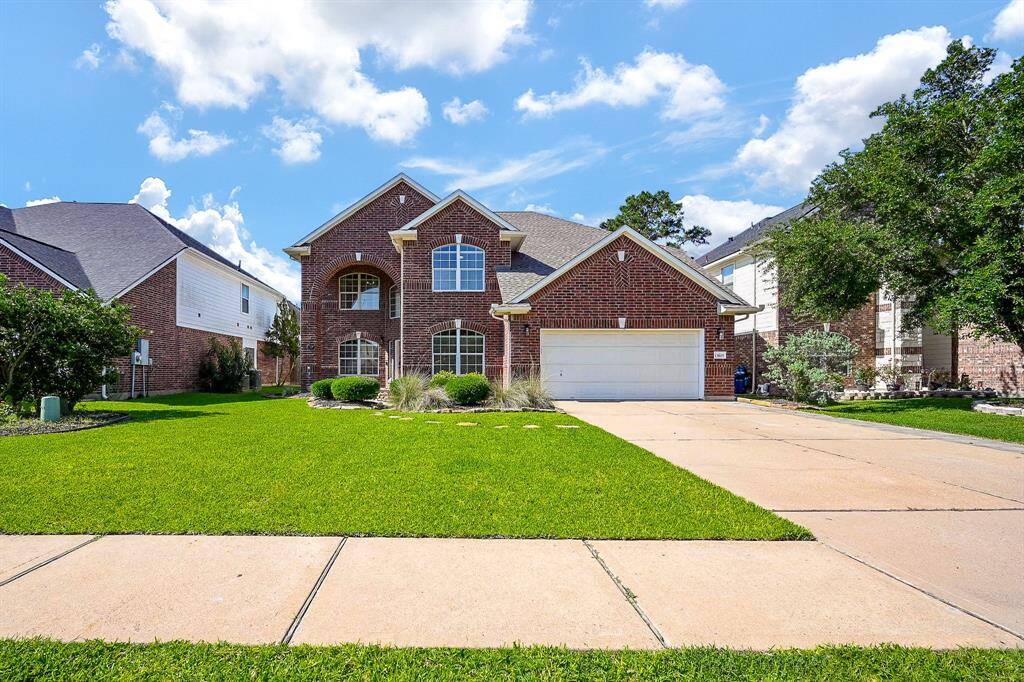 This photo showcases a two-story brick house with a well-maintained lawn and a spacious driveway leading to a two-car garage. The home features large windows and a welcoming entrance, set in a suburban neighborhood with similar styled homes.