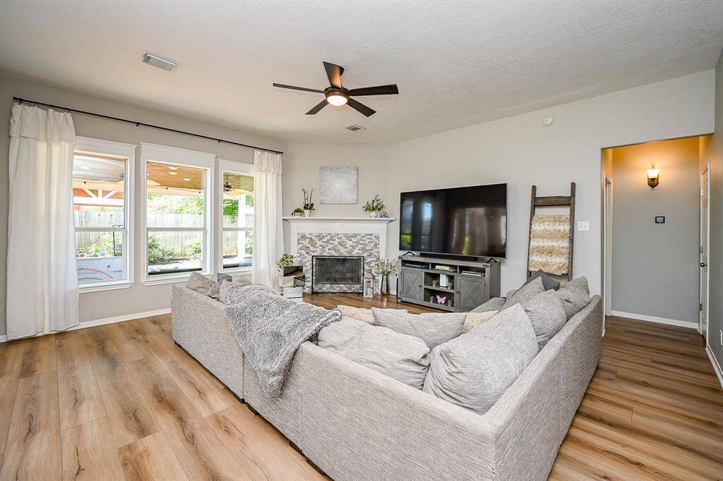 This inviting living room features plenty of space for cozy living are with a modern fireplace, and Natural light pours in through wide windows overlooking the backyard, while wide planked vinyl floors and neutral walls provide a warm, welcoming atmosphere.