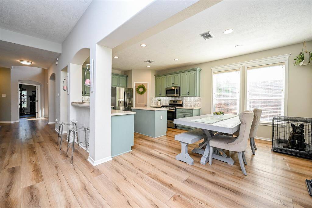 This photo showcases a bright, open-concept kitchen and dining area with light flooring. The kitchen features green cabinetry, stainless steel appliances, and a breakfast bar. Large windows provide ample natural light, and there is a cozy breakfast area.