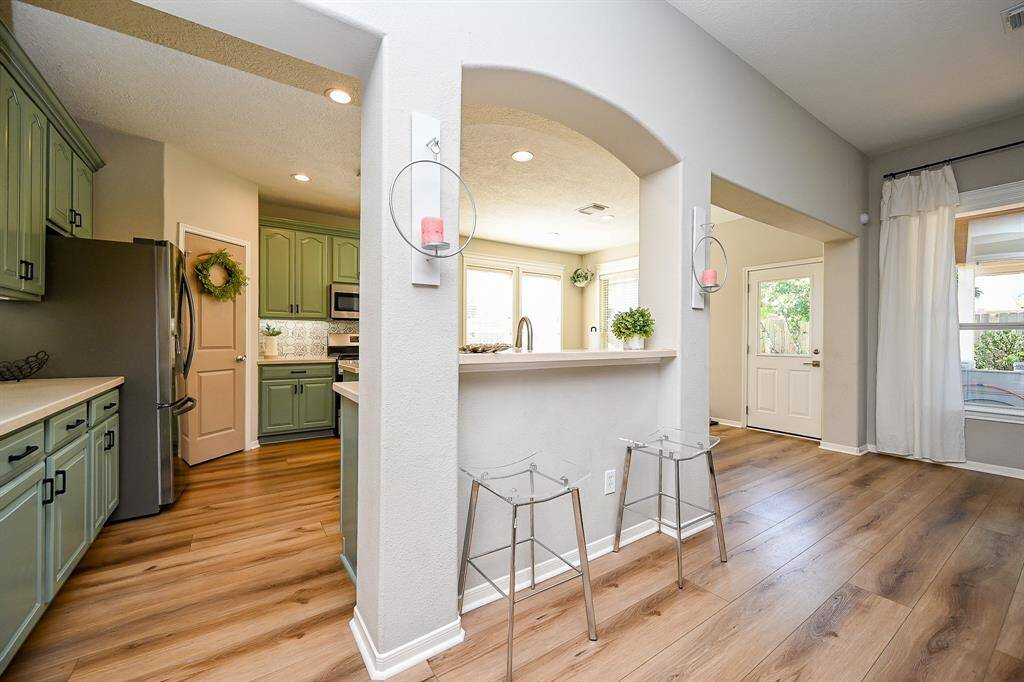 This photo showcases a bright, open kitchen with green cabinetry, stainless steel appliances, and vinyl flooring. An arched opening leads to a cozy living area with large windows and natural light. The space feels welcoming and well-lit, ideal for entertaining.