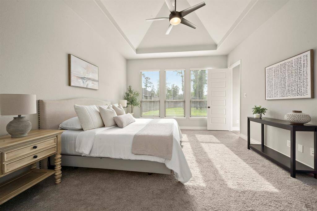 Discover a spacious, sunlit bedroom with a tray ceiling and large windows offering scenic views. The neutral decor, cozy bed, and modern ceiling fan create a serene, inviting retreat.