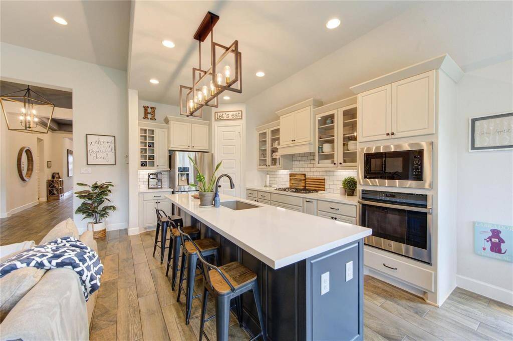 This modern kitchen boasts an open, airy feel overlooking the living area.  The large island/breakfast bar, Quartz countertops, sleek upgraded light fixture, and stainless steel appliances. compliment the white cabinets with glass fronts and subway tile backsplash. The space is bright and welcoming, ideal for entertaining.