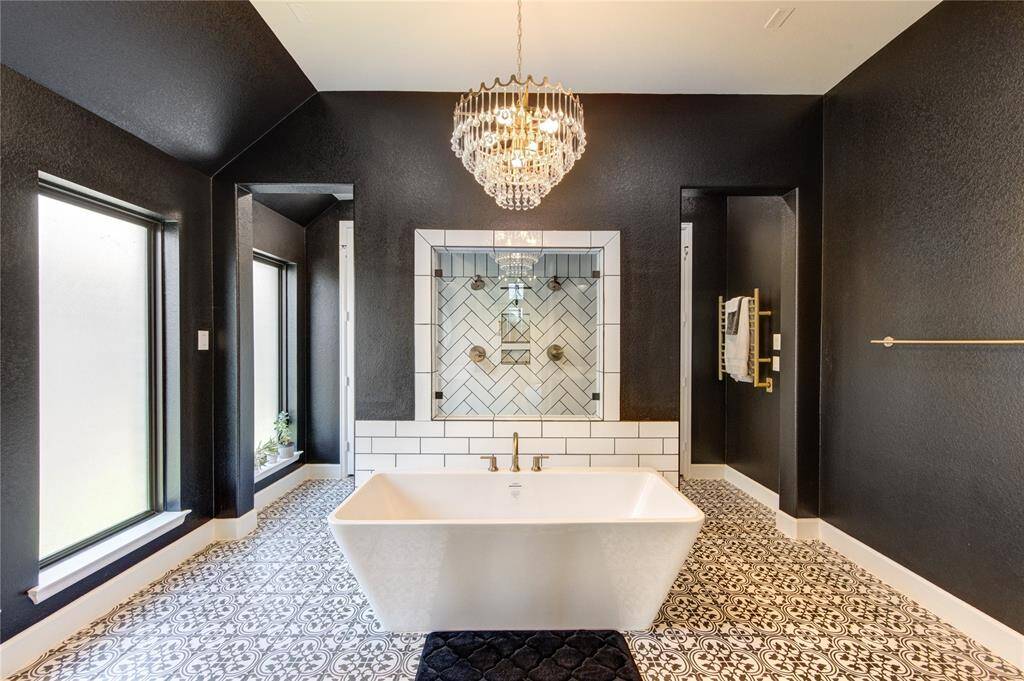 This well designed room features a luxurious freestanding tub, elegant chandelier, and stylish black and white patterned tile flooring. The space is accented by dark walls and contrasting white tiles, creating a modern and sophisticated atmosphere.