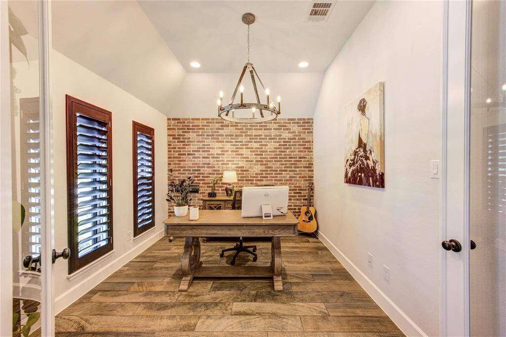 The charming home office features a rustic brick accent wall, stylish wood flooring, a chandelier, and plantation shutters, perfect for productivity and inspiration.
