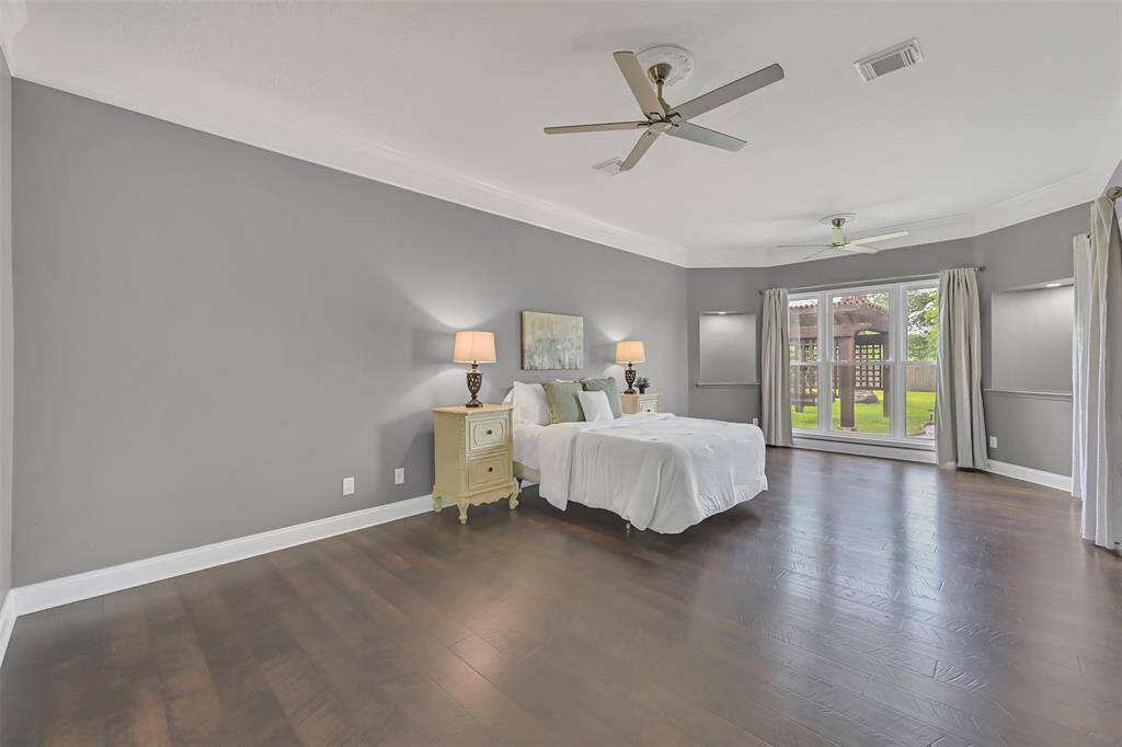 Expansive first-floor primary suite with rich Pergo wood floors, dual ceiling fans, custom wall niches, and oversized windows offering tranquil views of the backyard pergola and lush lawn—your private retreat awaits.