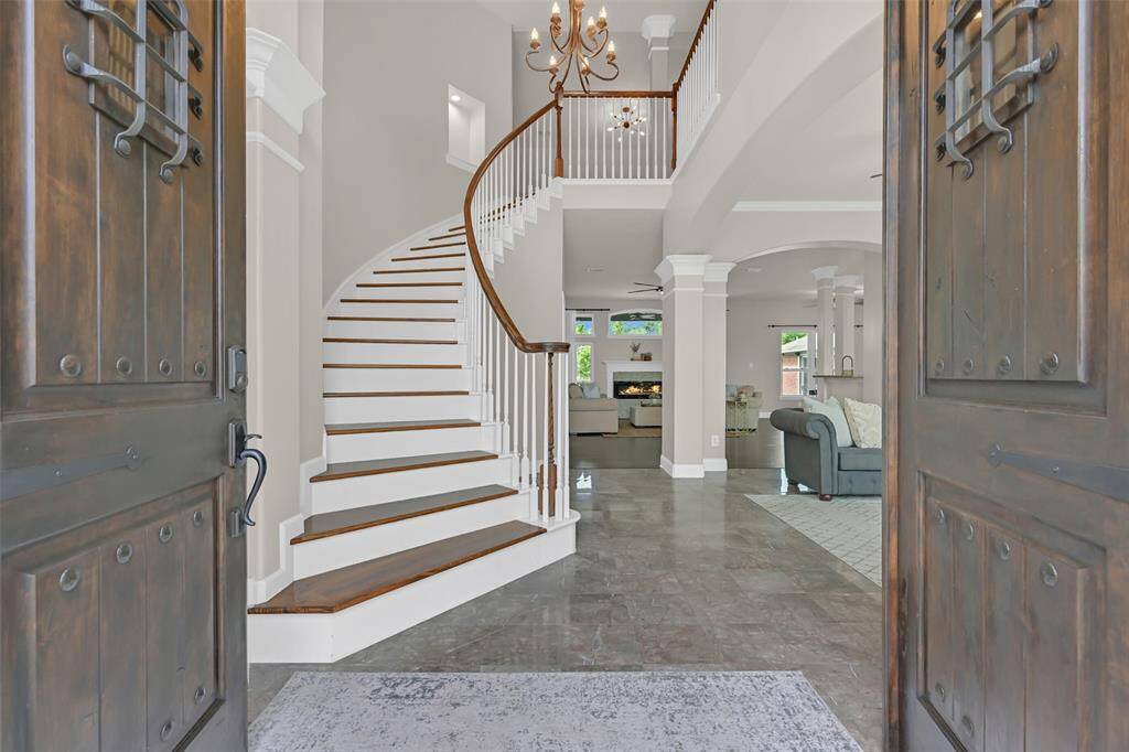 Step through solid wood double doors with speakeasy-style grilles & decorative clavos into a grand foyer with gleaming marble tile floors, soaring 19' ceilings, an elegant curved staircase, and columned archways that set the tone for refined living throughout.