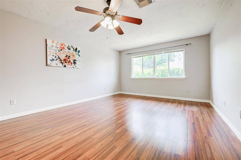 This primary bedroom features bright natural light from a large window, complemented by warm laminate flooring. It is spacious enough for a sitting area as well as all your bedroom furniture.