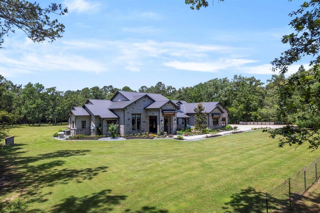 This stunning custom-built estate is beautifully set within a private, wooded 5-acre retreat. The home features a striking stone exterior, sleek metal rooflines, and meticulous landscaping that complements the lush, tree-lined backdrop. A wide front lawn leads to a grand entrance accented with elegant lighting and rich wood details, offering a warm welcome and a glimpse into the luxurious living within.
