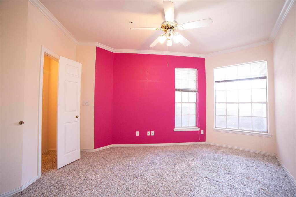This room features a bright accent wall, plush carpet, and ample natural light from two large windows. A ceiling fan adds comfort, and there's a nearby door leading to another space, enhancing its functionality.